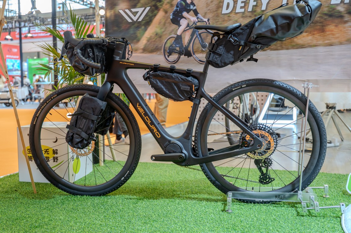Close-up of the Velduro Phantom bike featuring a sleek black design, equipped with bags for storage, displayed on grass-like flooring at an expo.