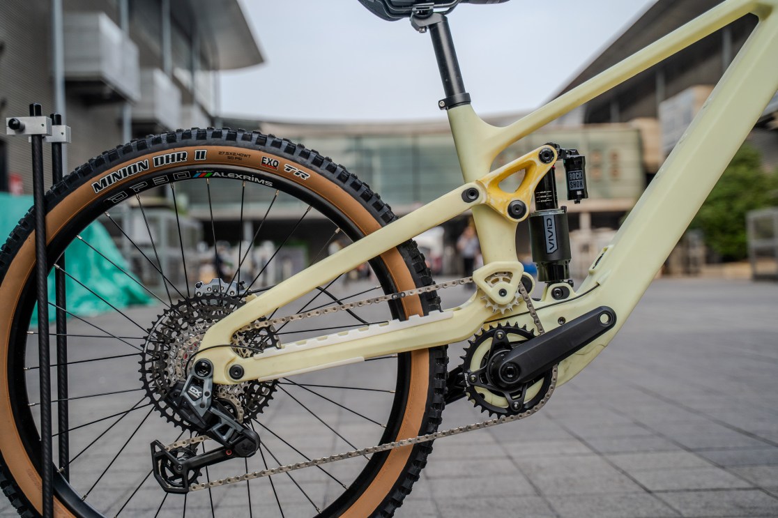 Close-up view of a sleek, light-colored mountain bike frame showcasing a mid-pivot suspension system and a powerful drivetrain with a black derailleur and a large cassette. The bike is equipped with Minion DHR II tires and has a stylish design, set against an outdoor expo backdrop.