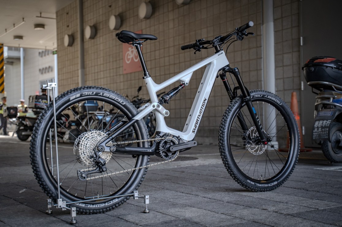 A Smartmotion Xplore carbon electric mountain bike displayed outdoors, featuring a sleek white frame, thick tires, and a mid-drive motor, with other bikes visible in the background.