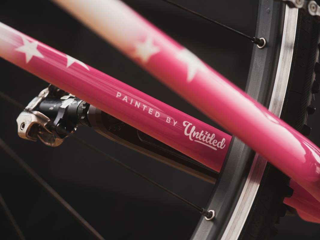Close-up of a bicycle frame showcasing a pink paint finish with white star patterns and the text 'PAINTED BY Untitled' on the downtube.