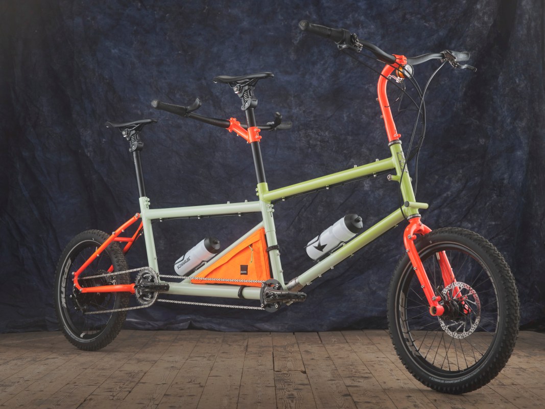A custom tandem bike featuring two seats, colorful frame components, and water bottle holders, set against a dark blue backdrop.