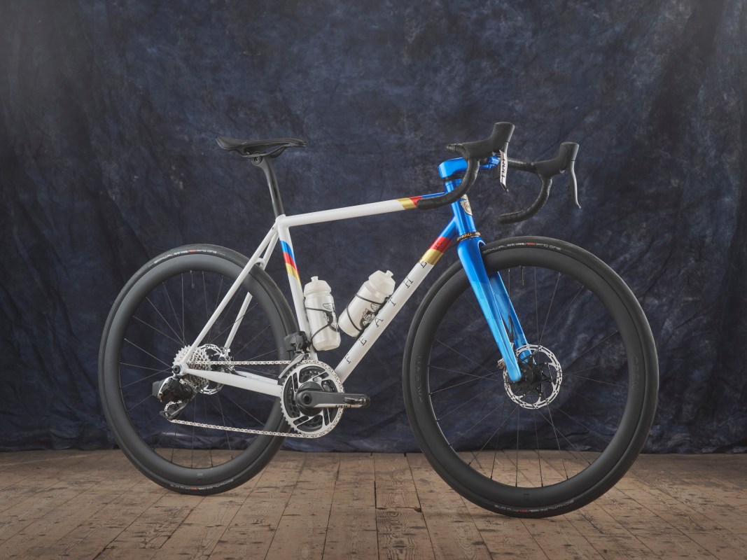 A custom-painted road bike featuring a white frame with vibrant geometric designs in blue, red, and yellow, placed on a wooden floor against a dark textured backdrop.