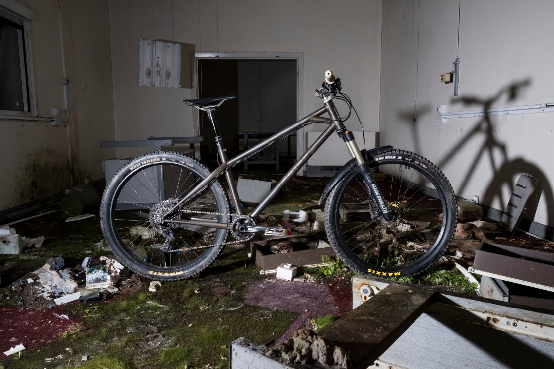 A custom hardtail mountain bike displayed in an abandoned room, surrounded by debris and overgrown moss.