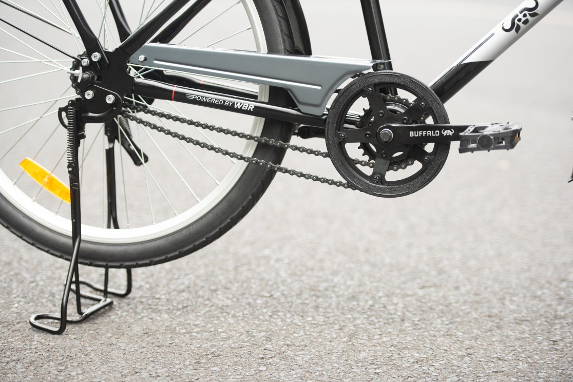 Close-up of a bicycle's rear drive train, showcasing the black crankset, chain, and pedal alongside a kickstand.