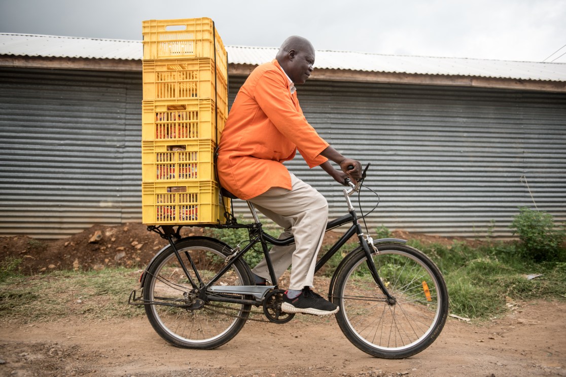 A person wearing an orange shirt rides a Buffalo Bike S2 loaded with yellow crates. The bicycle is on a dirt path with greenery and a corrugated metal wall in the background.