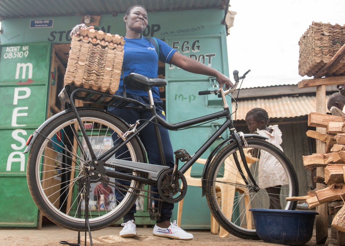 A young woman stands proudly next to a black Buffalo Bike S2, holding a stack of egg cartons on the rear rack, with a shop displaying 'mpesa' in the background.