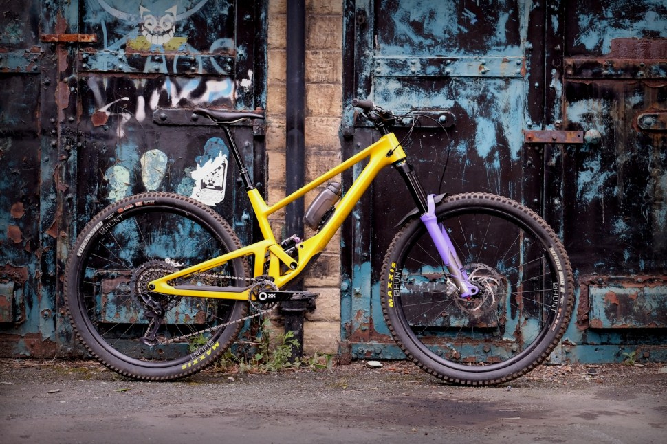 A bright yellow mountain bike with purple accents parked against a grungy blue and rusty wall.