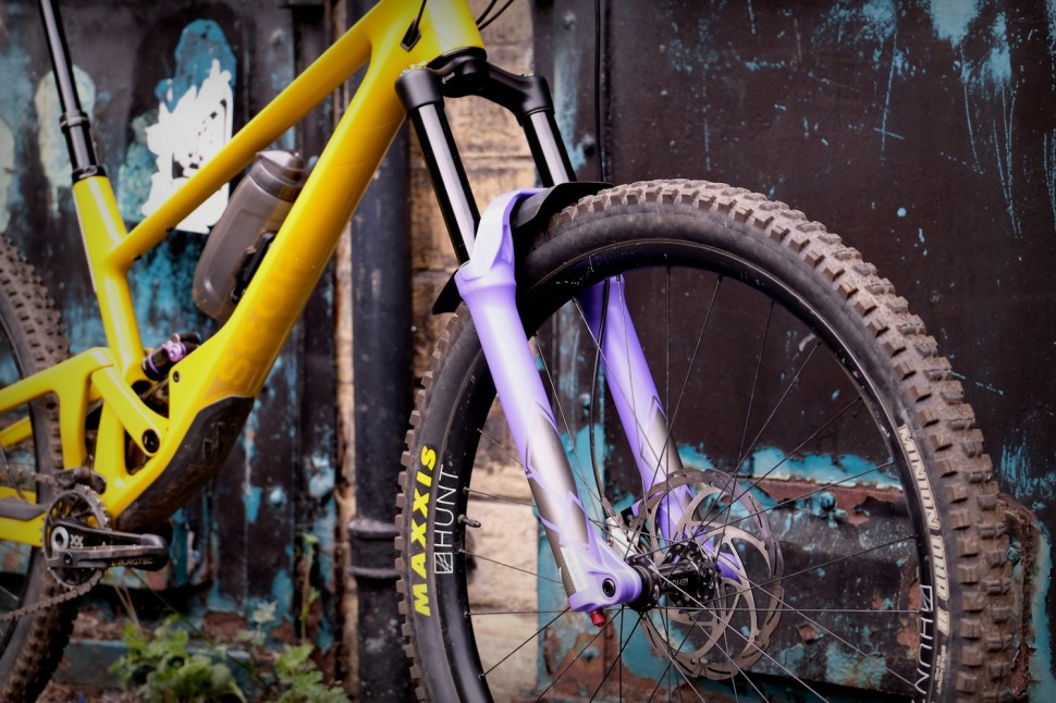 Close-up of a yellow mountain bike frame with a purple and silver fork, showcasing the tire and brake disc against a textured, weathered background.