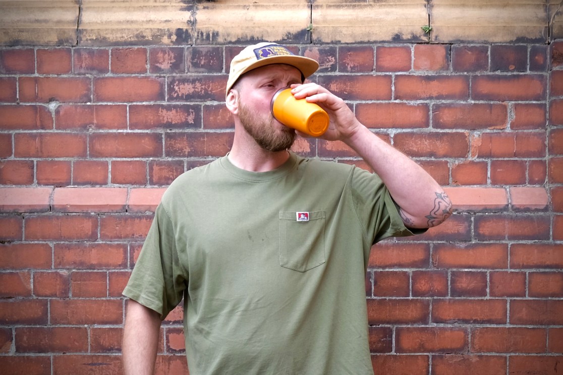 A man wearing an olive green t-shirt and a baseball cap drinks from an orange cup while standing in front of a brick wall.