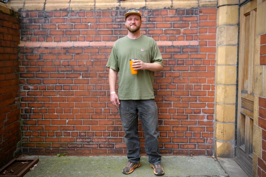 A person standing against a brick wall, wearing a green t-shirt and holding a drink in an orange cup.