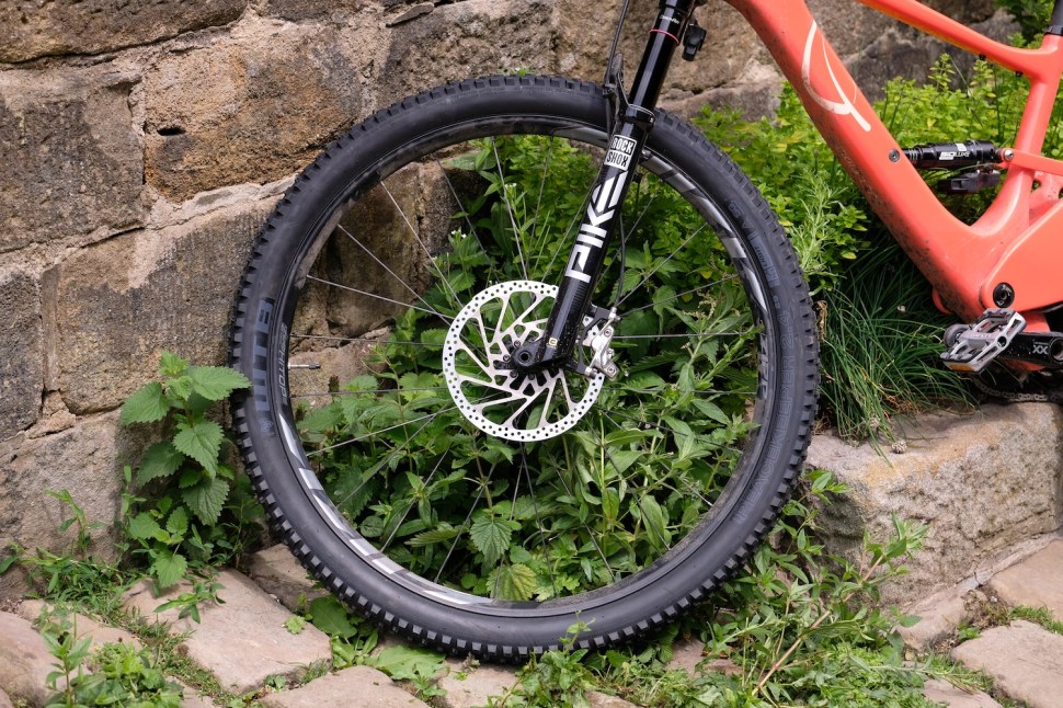 Close-up of a mountain bike wheel with a disc brake, resting on grass and cobblestone pavement.