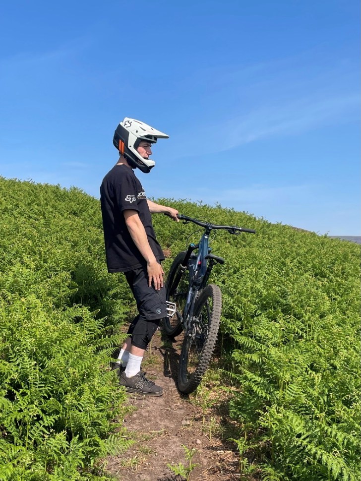 A mountain biker standing next to a bike on a path surrounded by green ferns under a clear blue sky, wearing a white full face helmet.