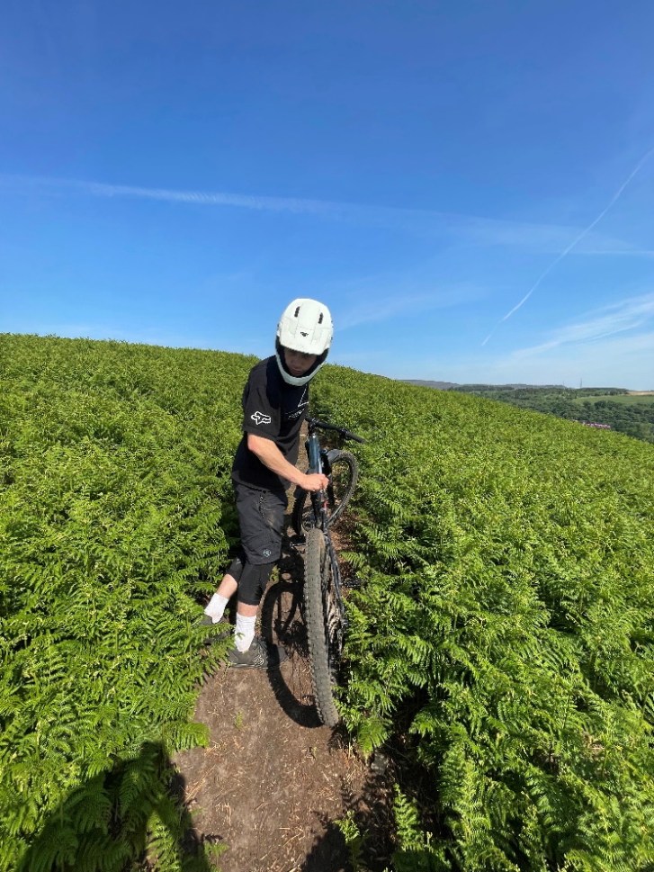 A person wearing a white full face mountain bike helmet poses with their bike on a trail surrounded by green ferns under a blue sky.