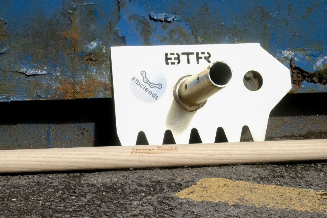 A close-up of a white bike trail tool with the letters 'BTR' engraved on it, resting on a wooden handle, against a textured blue and rust-colored background.