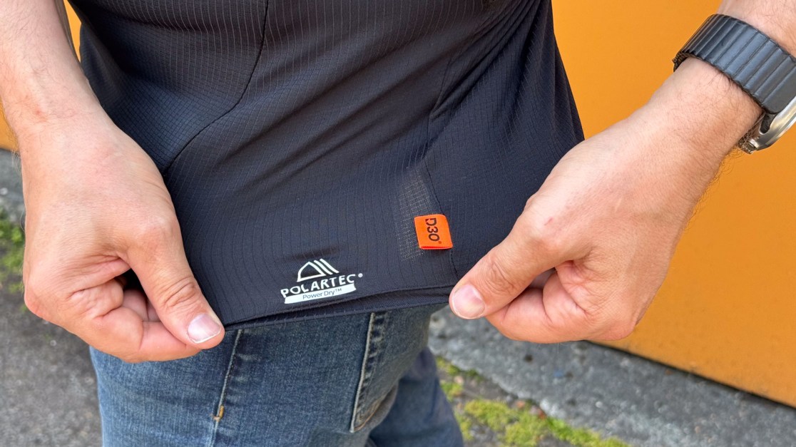 Close-up of a person's hand holding the hem of a black sports shirt, showcasing the Polartec label for Power Dry fabric.