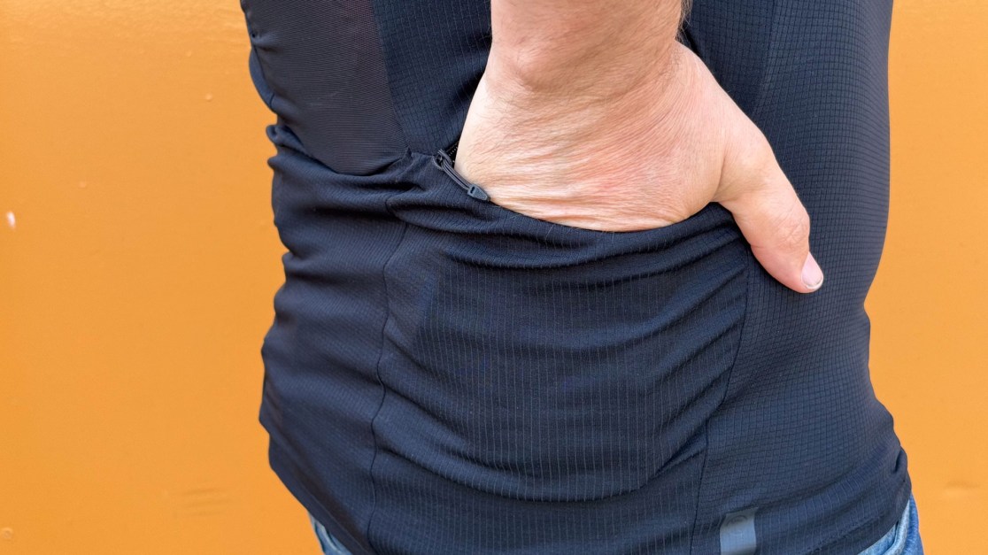 Close-up of a person's hand reaching into the side pocket of a fitted black shirt against an orange background.