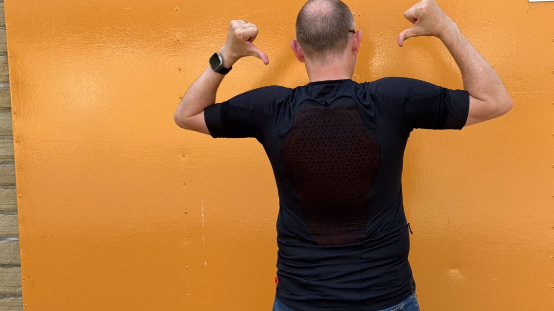 A person standing against an orange wall, showing off a black jersey with a patterned back while giving a thumbs-up.