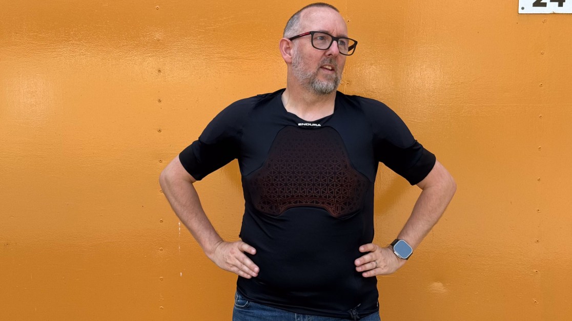 A person standing against an orange wall, wearing a fitted black protective shirt with a geometric padding design on the chest. The individual has short hair and glasses, and is posing with hands on hips.