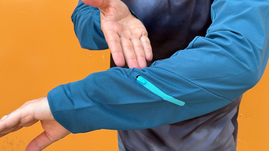 A close-up of a person's arm wearing a blue long-sleeve shirt, showcasing a zippered pocket on the forearm against an orange background.