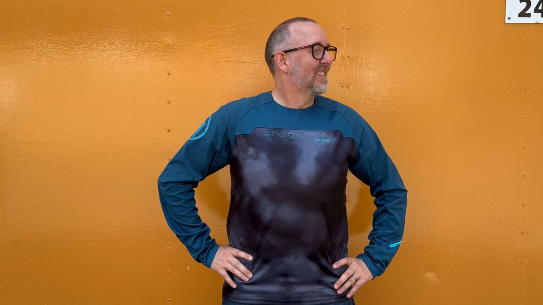 A man wearing a long-sleeve sporty jersey with 'ENDURA MT500' branding, standing against an orange background.