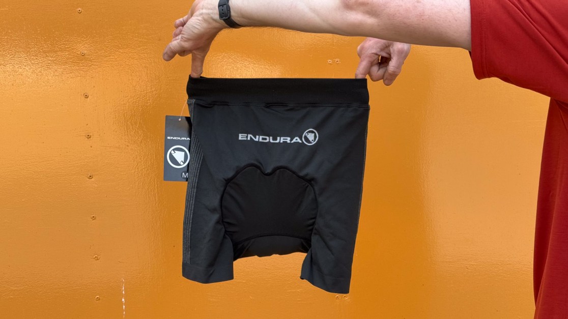 A person holding up a pair of black Endura cycling shorts in front of an orange background.