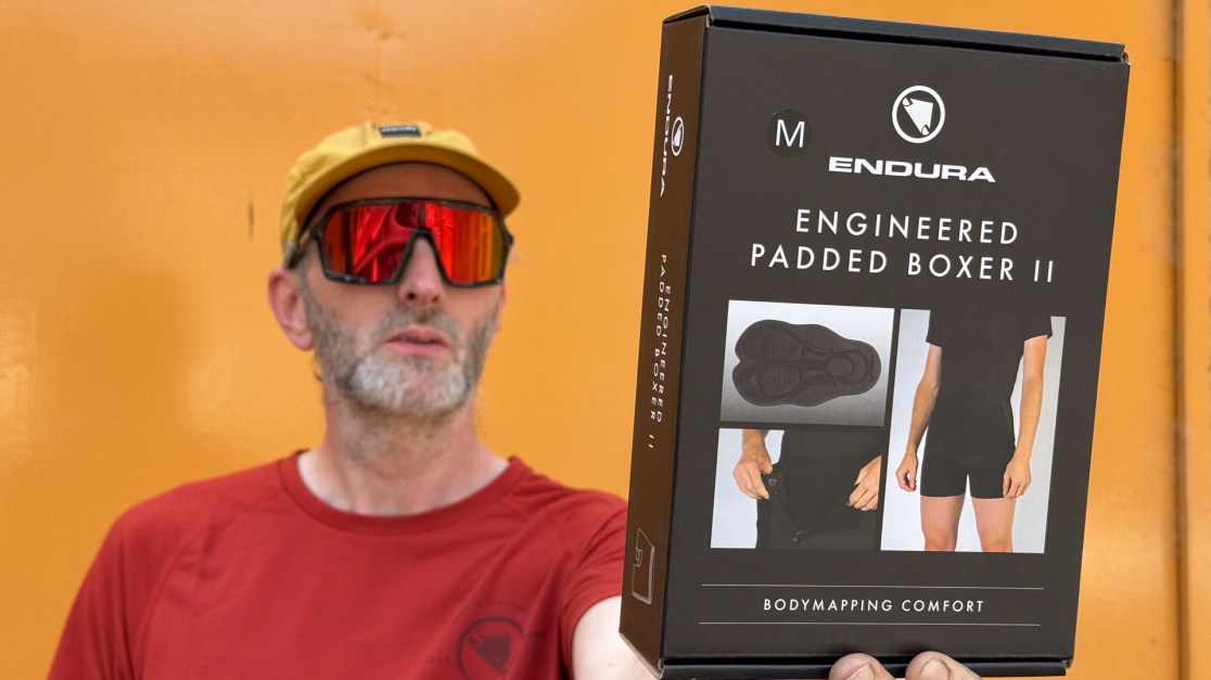 A person holding a box of Endura Engineered Padded Boxer II against an orange background, wearing a yellow cap and red mirrored sunglasses.