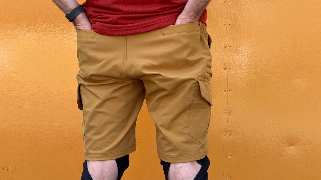 Person wearing brown cargo shorts and a red shirt, standing in front of an orange background.