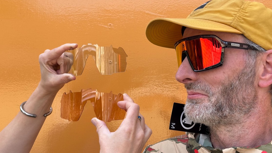 A person wearing bright sunglasses holds up a clear replacement lens while looking at another individual. The background is painted in an orange color.