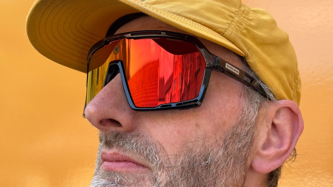 Close-up of a man wearing a yellow cap and large reflective sunglasses, with a focus on his facial features against an orange background.
