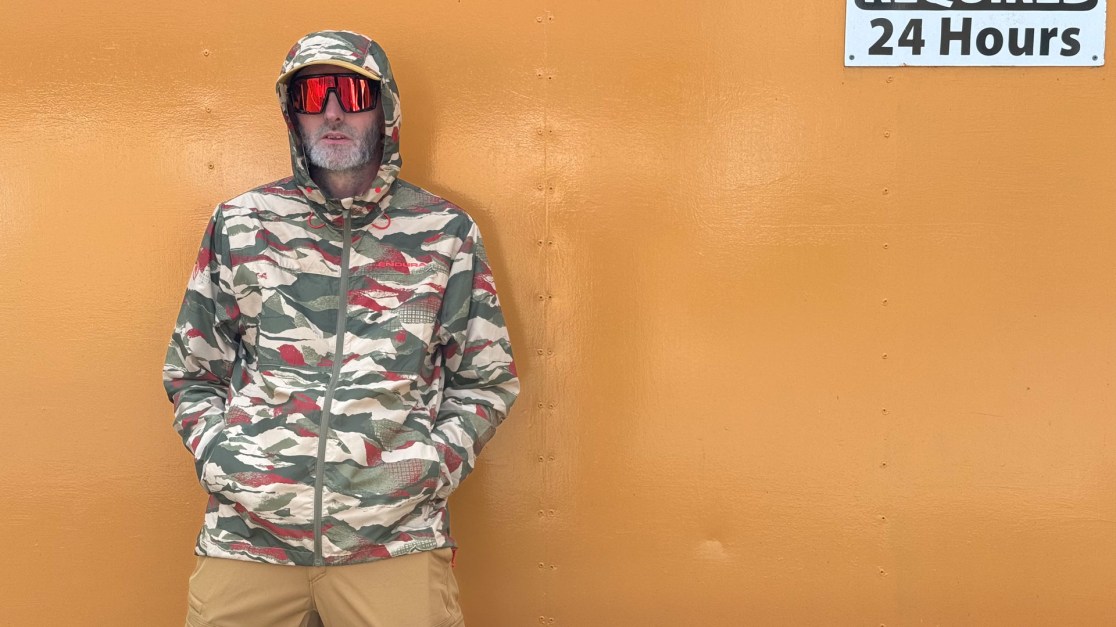 A person wearing a camouflaged jacket and sunglasses stands against a bright orange background.