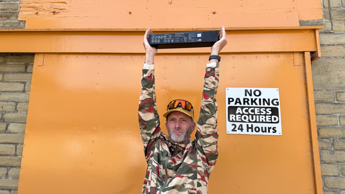 A man in a camouflage jacket holding a rectangular object above his head in front of an orange wall with a sign that reads 'NO PARKING ACCESS REQUIRED 24 HOURS'.