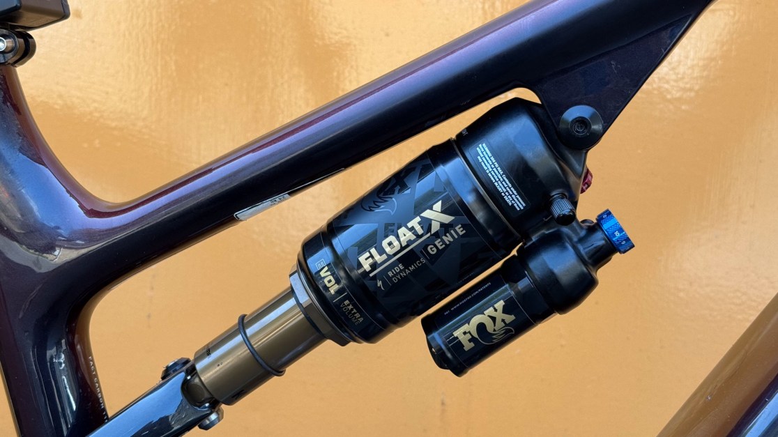 Close-up view of the FOX FLOAT X shock absorber mounted on a mountain bike frame, showcasing details like the adjustment knobs and branding against a solid orange background.