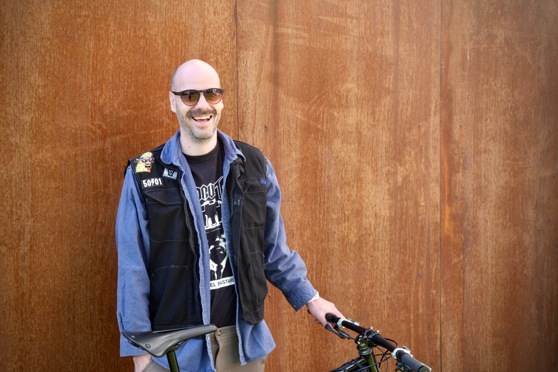 A smiling person wearing sunglasses and a vest stands beside a bike, with a rustic metal background.