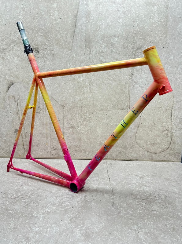 A vibrant, multicolored bike frame displaying an orange to pink gradient, with the word 'FEATHER' painted along the side.