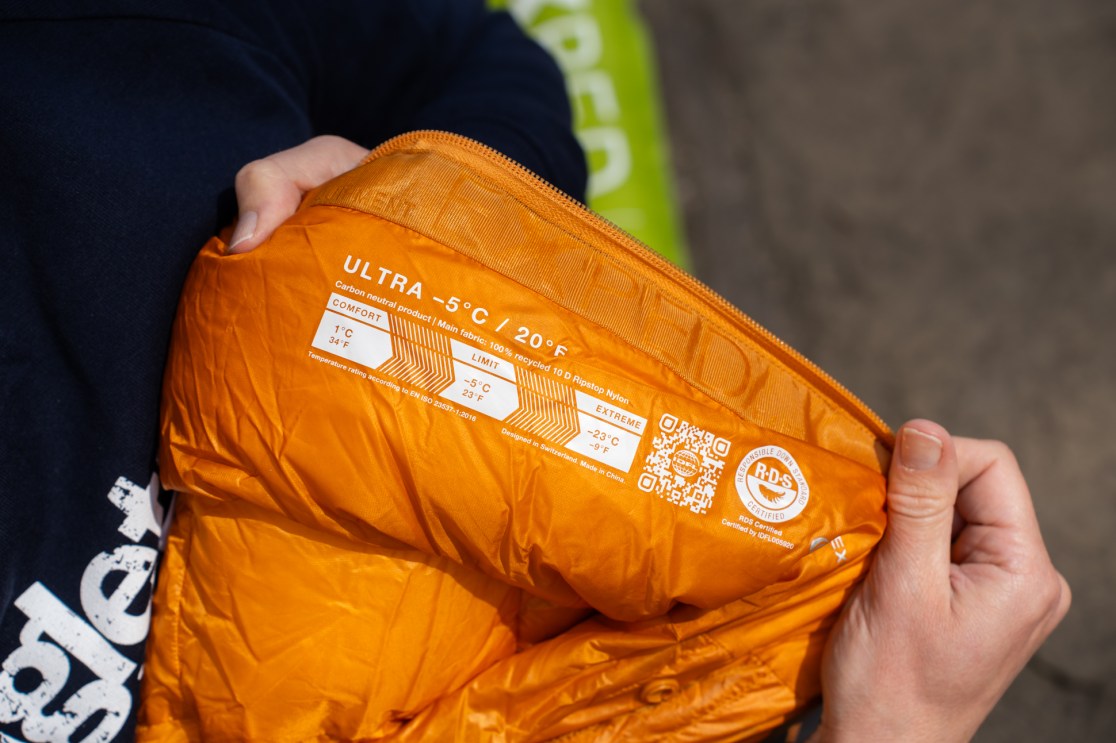Close-up of an orange sleeping bag showing temperature ratings and materials, held by a person in a dark sweater.