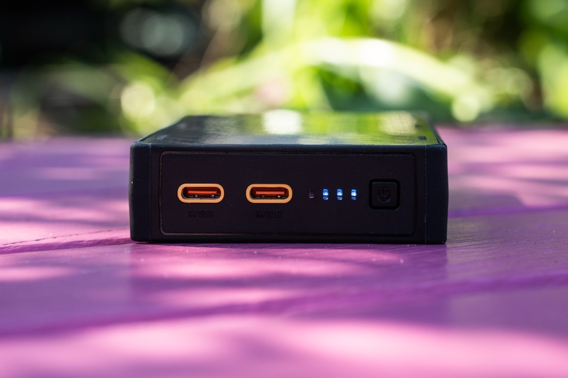 Close-up view of a portable battery pack with two USB-C ports and indicator lights, placed on a pink surface with blurred greenery in the background.