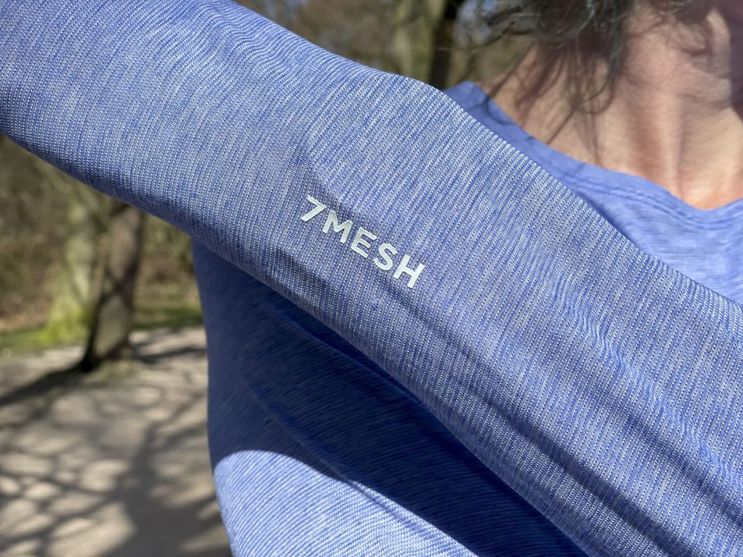 Close-up of a person's arm wearing a blue athletic shirt featuring the '7MESH' logo, surrounded by a natural outdoor setting.