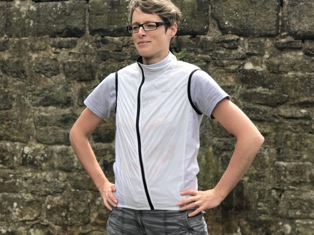 A person stands confidently with their hands on their hips, wearing a lightweight, windproof gilet over a gray t-shirt, in front of a stone wall.