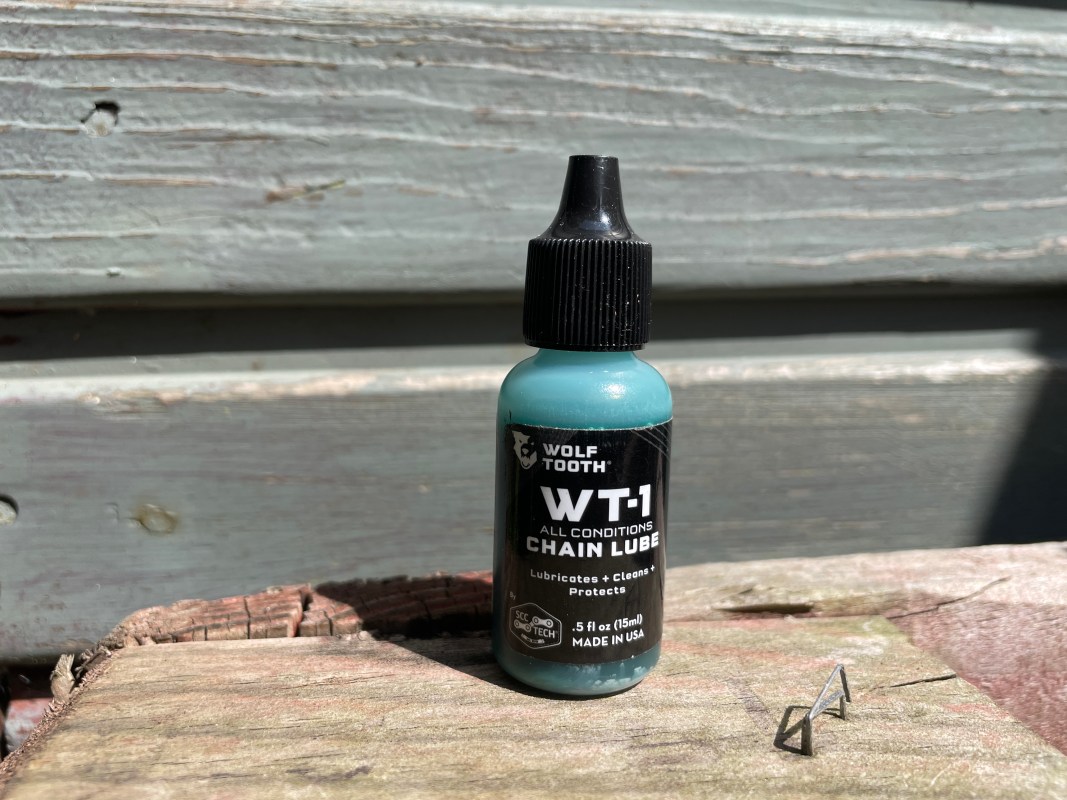 A bottle of Wolf Tooth WT-1 all conditions chain lube, placed on a wooden surface with weathered paint, next to small metal chain connectors.