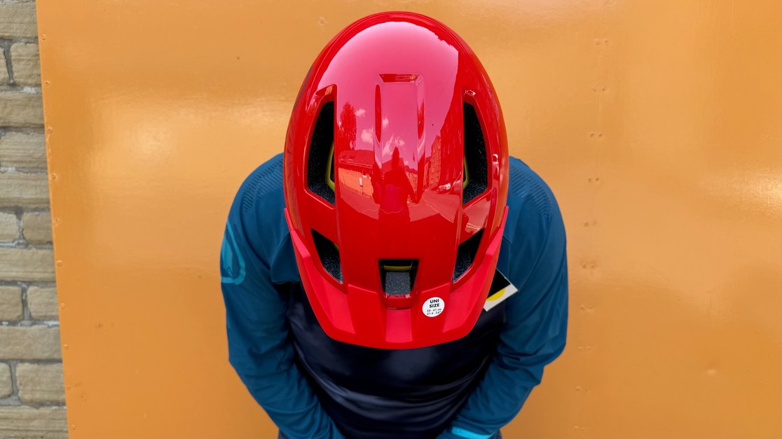 A red bike helmet viewed from above, resting on the head of a person wearing a blue long-sleeve shirt, with an orange background.