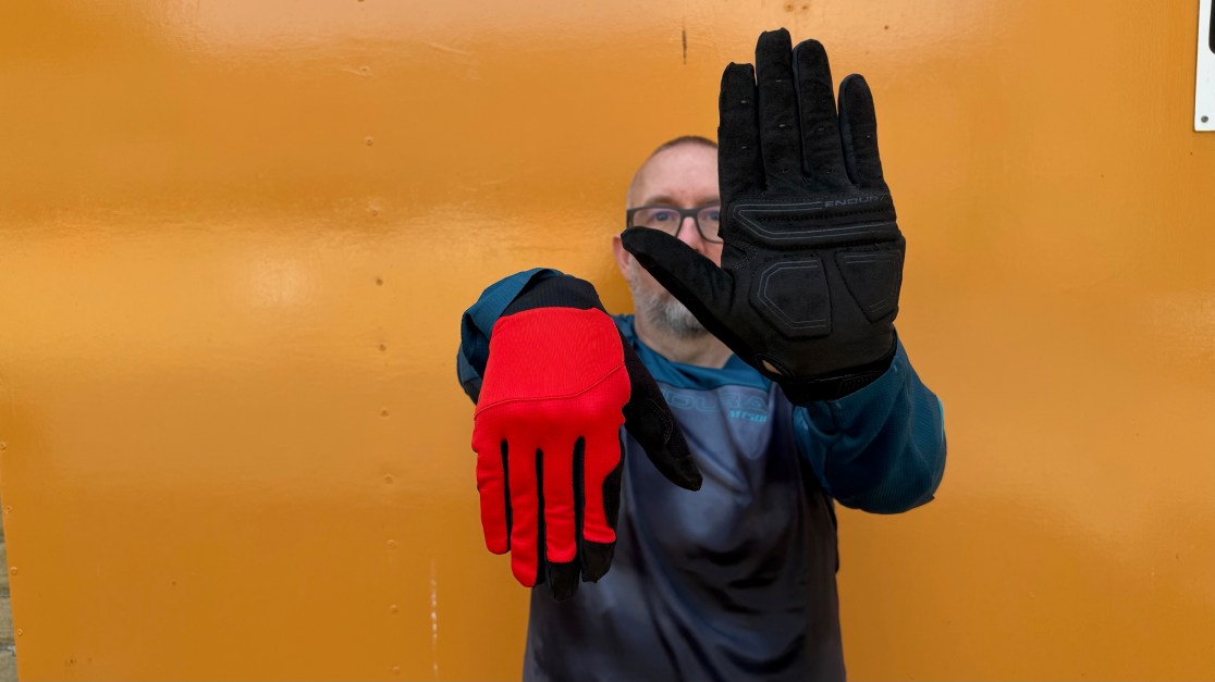 A person holding up two different styles of cycling gloves, one red and one black, against an orange background.