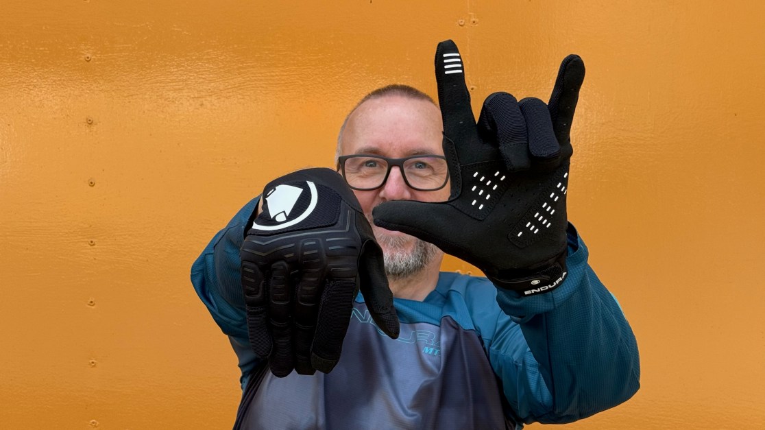 A person holding up two different cycling gloves in front of an orange background, showing enthusiasm.