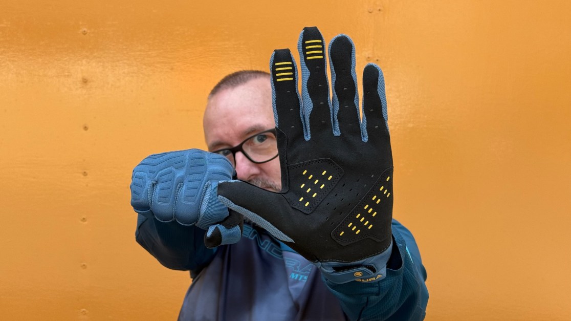 A person showing a pair of Endura SingleTrack gloves, one glove facing towards the camera and the other with fingers curled into a fist, in front of a bright orange background.
