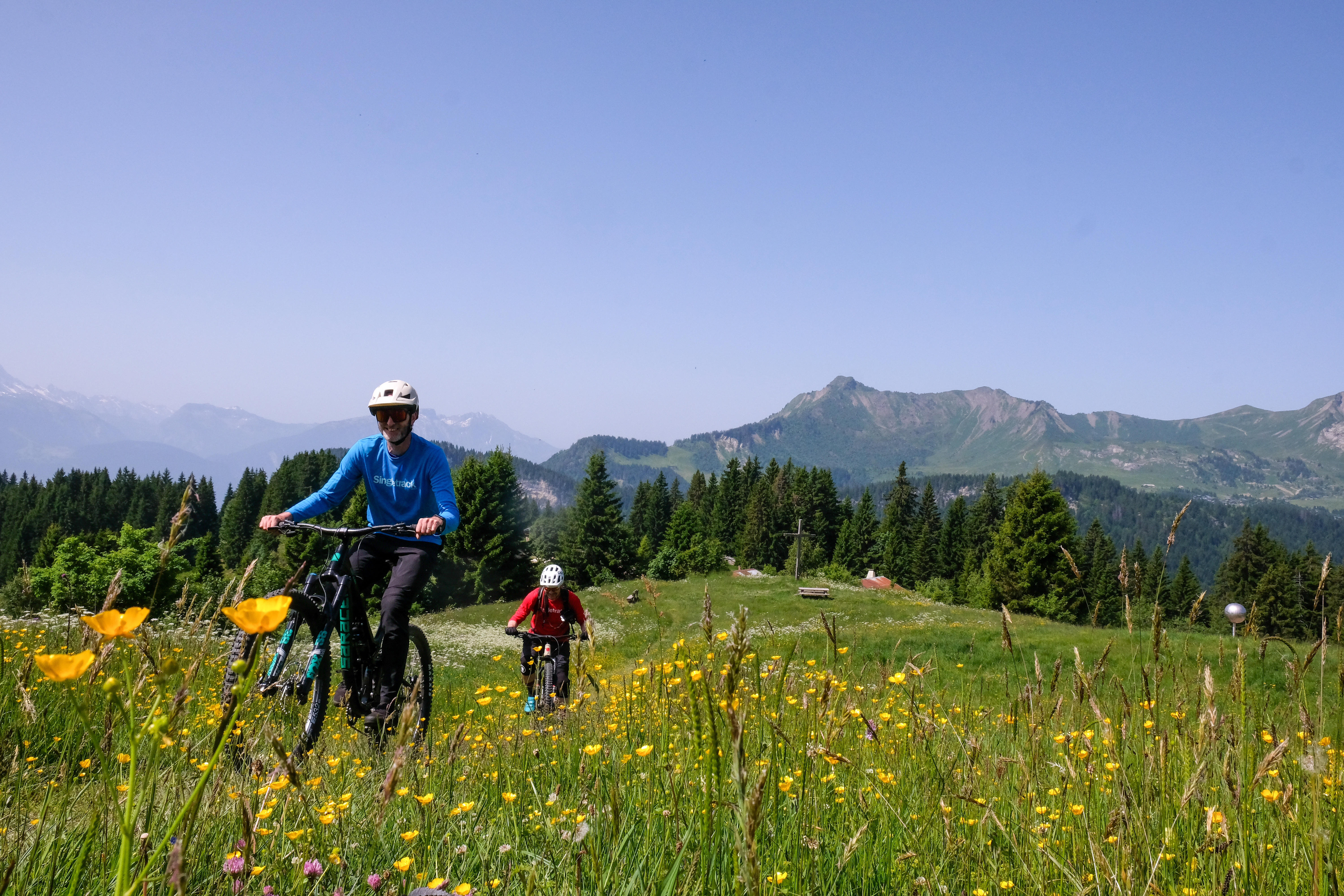 les gets porte du soleil destination guide two riders in singletrack shirts riding through a meadow of flowers with mountains in the background