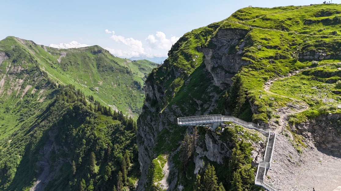 A panoramic view of the French Alps featuring lush green hills, rocky cliffs, and a display path winding along the edge of a mountain.