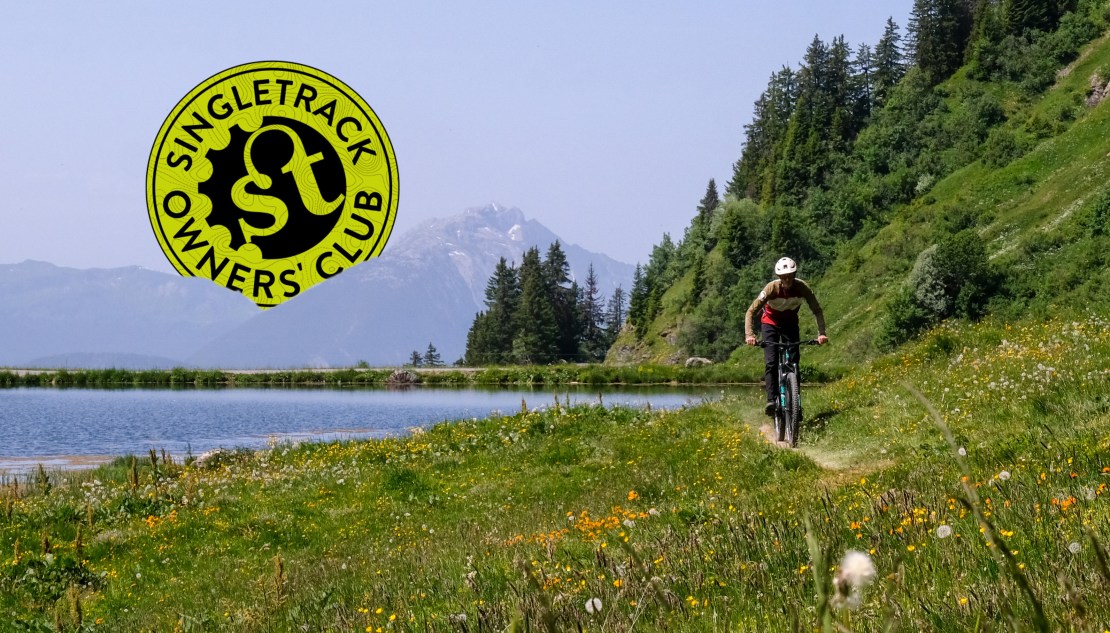 A mountain biker riding along a trail near a lake with lush greenery and flowers, with the Singletrack Owners' Club logo overlayed in the top left corner.