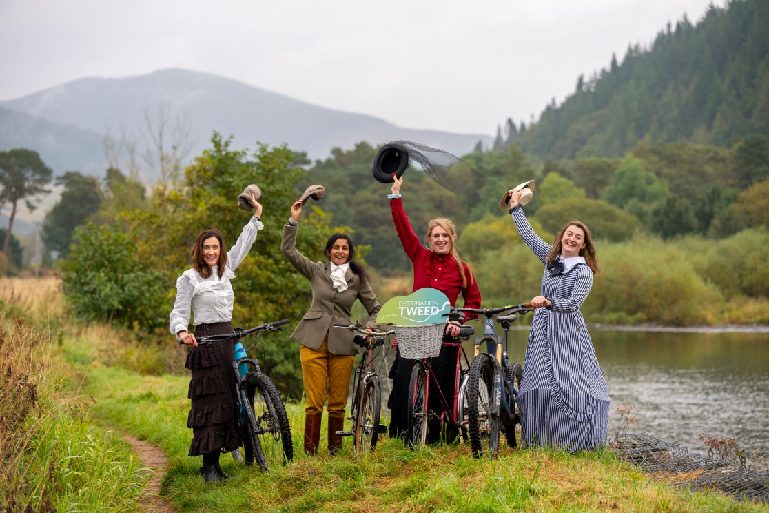 Four women in historical outfits stand by bicycles near a river in Tweed Valley, raising their hats in celebration.