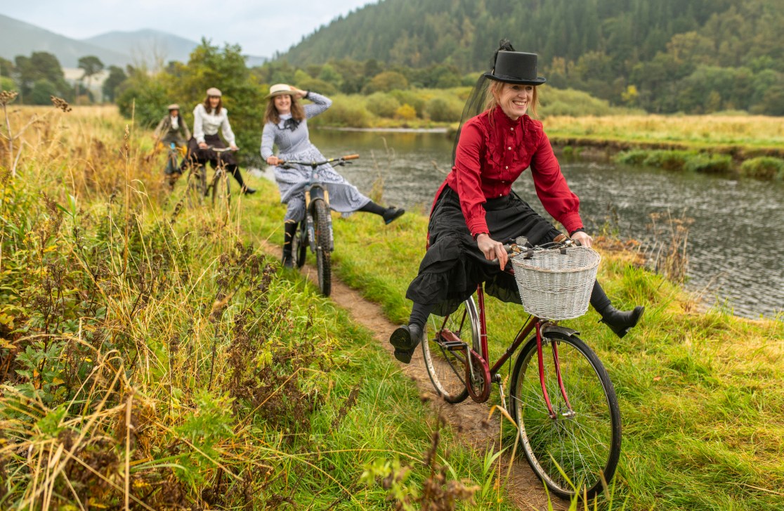 A group of women in vintage attire riding bicycles along a scenic path beside a river in the Tweed Valley, Scotland.