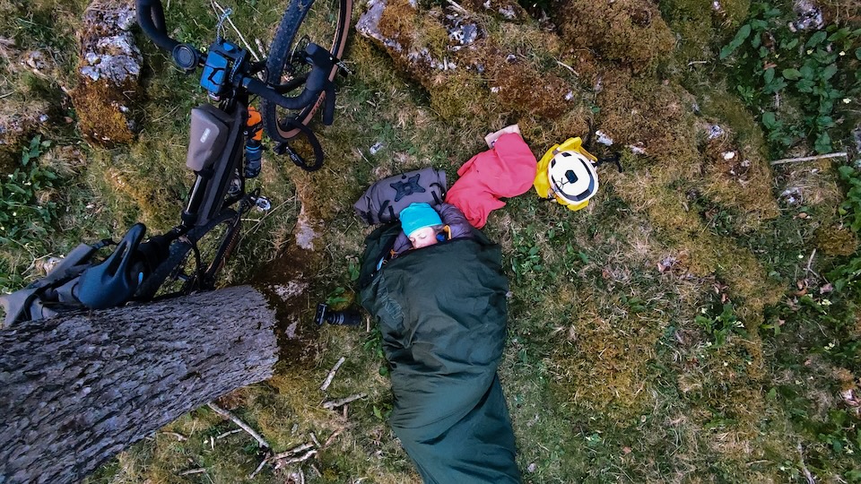 A person sleeping peacefully in a sleeping bag under a tree, with a bicycle and camping gear nearby, surrounded by green grass and moss.