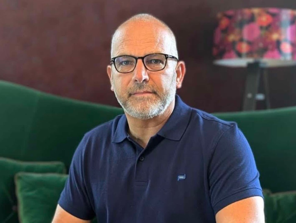 A middle-aged man with glasses and a short beard, wearing a navy polo shirt, sits in a green sofa setting with a colorful lamp in the background.